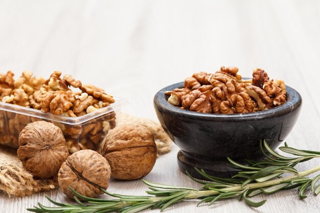 Peeled and unpeeled walnuts in stone bowl and plastic container with sackcloth and rosemary on a wooden background. Useful nutritious protein product. Shallow depth of field.