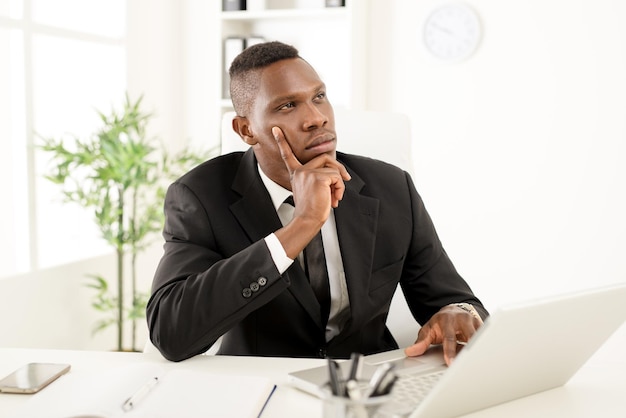 Peinzende Afrikaanse zakenman die op kantoor zit en op een laptop werkt. Wegkijken.