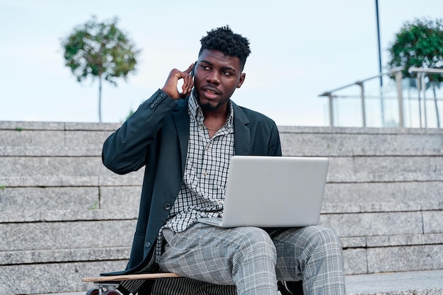 Peinzende Afro-Amerikaanse student in colbert zittend met laptop open op stenen trappen in de straat en pratend op smartphone bij daglicht