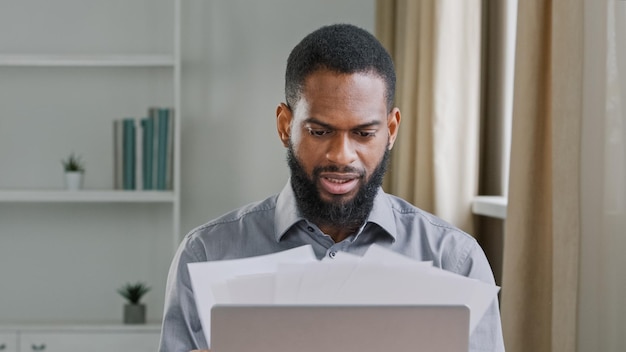 Peinzende duizendjarige etnische bebaarde mannelijke kantoormedewerker die papierwerk analyseert ernstige afro-amerikaanse zakenman leider man werkgever werken met papieren documenten rapporteren gegevensinformatie op de werkplek