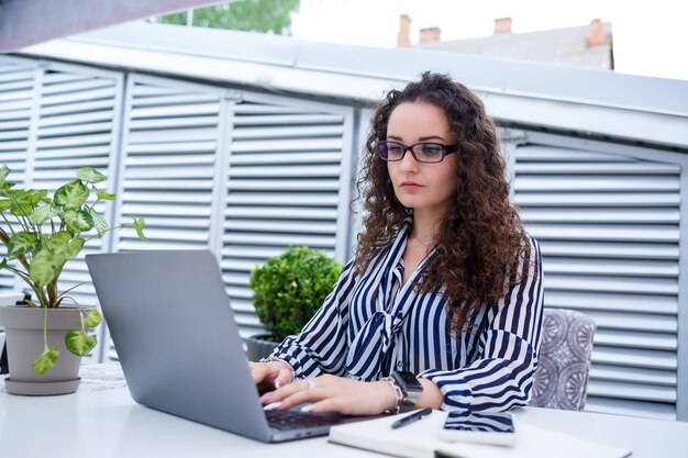 Peinzende freelancer-vrouw in trendy vrijetijdskleding die met een laptop werkt en aantekeningen maakt, lacht en kijkt naar het terras van het café