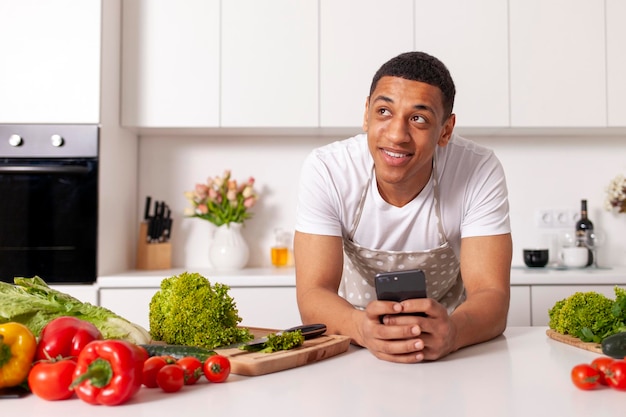 Peinzende jonge Afro-Amerikaanse man in schort gebruikt smartphone in witte moderne keuken en dromen