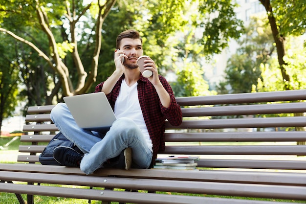 Peinzende jonge man praten op mobiel, werken op laptop en koffie drinken, zittend in park op bankje. Technologie, communicatie, onderwijs en werken op afstand concept, kopieer ruimte