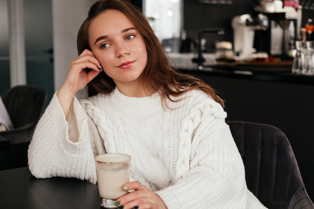 Peinzende jonge vrouw die binnen koffie drinkt Kaukasisch meisje zit in café in trui