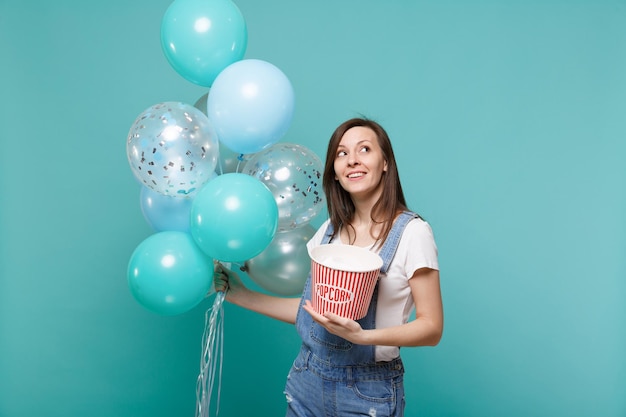 Peinzende jonge vrouw in denim kleding opzoeken met emmer popcorn vieren met kleurrijke lucht ballonnen geïsoleerd op blauwe turkooizen achtergrond. Vakantie verjaardagsfeestje, mensen emoties concept.