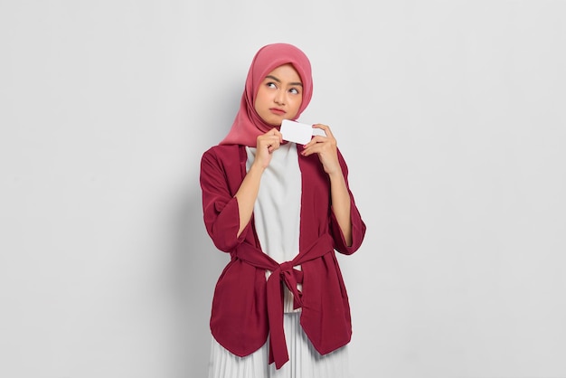 Peinzende mooie Aziatische vrouw in een casual shirt en hijab met een creditcard, kijkend naar lege ruimte geïsoleerd op een witte achtergrond. Mensen religieuze levensstijl concept