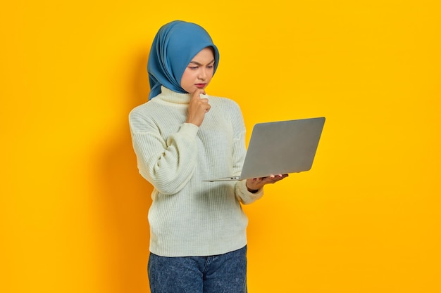 Peinzende mooie Aziatische vrouw in witte trui met laptop kijkt serieus na over iets geïsoleerd over gele achtergrond mensen religieuze levensstijl concept