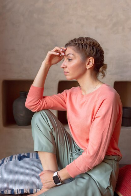 Peinzende mooie vrouw in koraal sweatshirt en grijze broek zit in de kamer