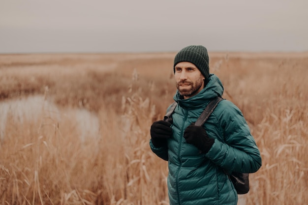 Peinzende ongeschoren man draagt hoed groene jas en handschoenen draagt rugzak poses tegen gele herfst veld achtergrond heeft een wandeling naar buiten ademt frisse lucht gefocust op afstand Toerisme concept