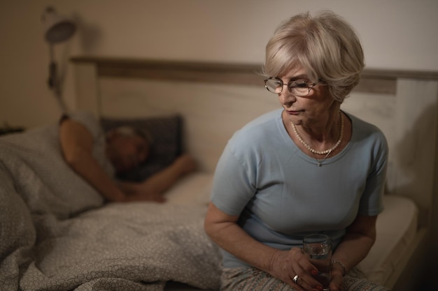 Peinzende rijpe vrouw die op het bed zit en aan iets denkt terwijl ze een glas water vasthoudt Haar man slaapt op de achtergrond