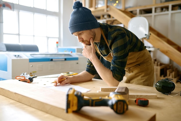 Peinzende slimme timmermansontwerper die naar een planschema kijkt dat over de werktafel leunt in een moderne werkplaats