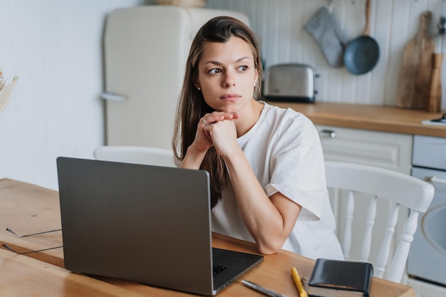 Peinzende Spaanse jonge volwassen vrouw zit aan tafel met laptop in keuken uitgeput van hard werken Verdrietig Italiaans meisje voelt vermoeidheid kijkt opzij denkt aan financiële problemen Overbelast vrouwelijk falen