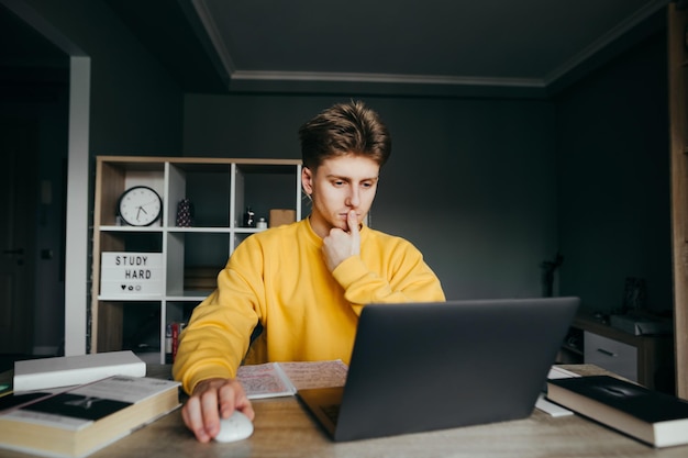 Peinzende student die thuis aan een tafel met boeken zit en een laptop gebruikt met een serieus gezicht Slimme tiener leert op afstand online tijdens quarantaine