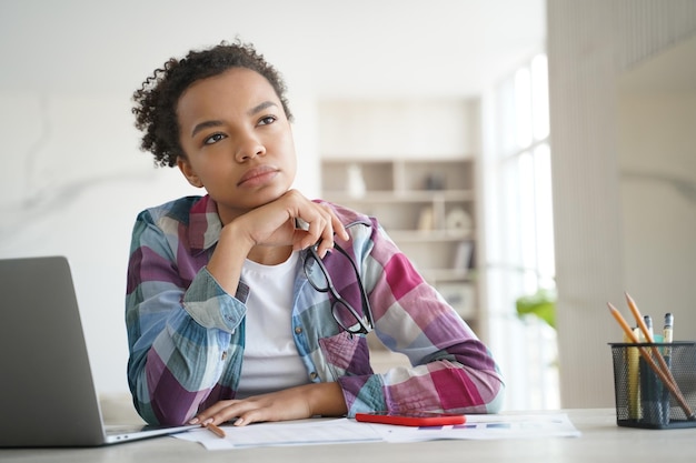 Peinzende tienermeisjesstudent van gemengd ras kijkt opzij, in gedachten verzonken terwijl hij thuis leert