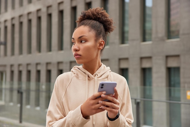 Peinzende vrouwelijke trainer gekleed in hoodie gebruikt smartphone voor online berichten geeft sportief advies heeft pauze tijdens training downloads muziek voor actieve training poses tegen stad gebouw achtergrond