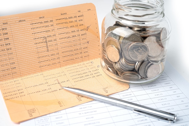 Pen on account book bank with coins in glass jar.