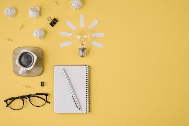 Pen, notepad, eyeglasses, cup of coffee, light bulb on yellow background.