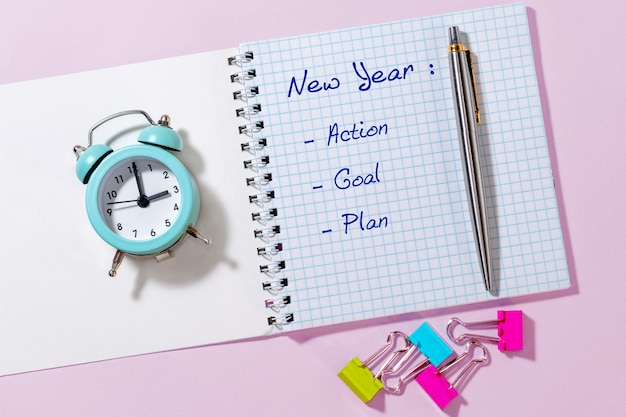Pen, papier met nieuwjaar, actie, doel, plan en blauwe wekker op lichtroze