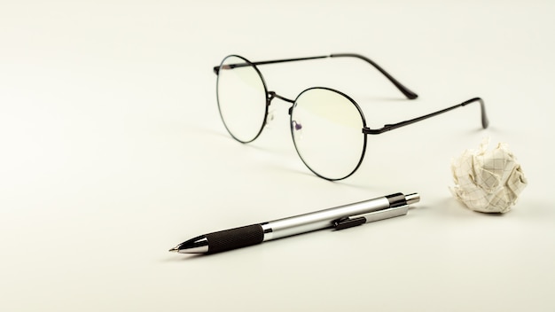 Pen with glasses and crumpled paper ball on white desk background.