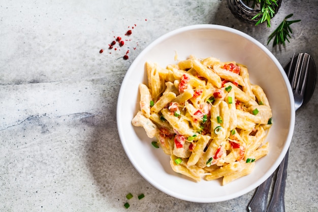 Penne pasta with chicken, pepper and green onions in creamy sauce in a white plate, gray background, top view.