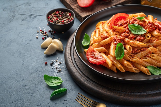 Penne pasta with tomato and meat bolognese sauce and fresh basil in a black plate on a dark background. Side view, close-up.