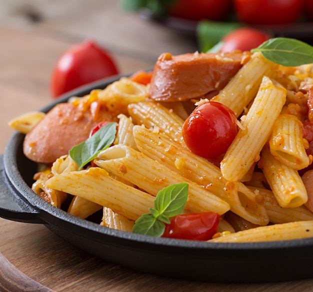 Penne pasta with tomato sauce with sausage, tomatoes, green basil decorated in a frying pan on a wooden table