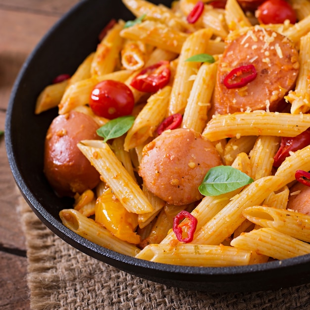 Penne pasta with tomato sauce with sausage, tomatoes, green basil decorated in a frying pan on a wooden table