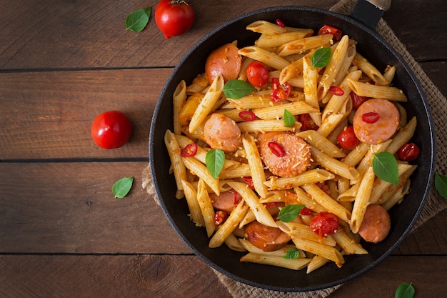 Penne al sugo di pomodoro con salsiccia, pomodori, basilico verde decorato in padella su un tavolo di legno