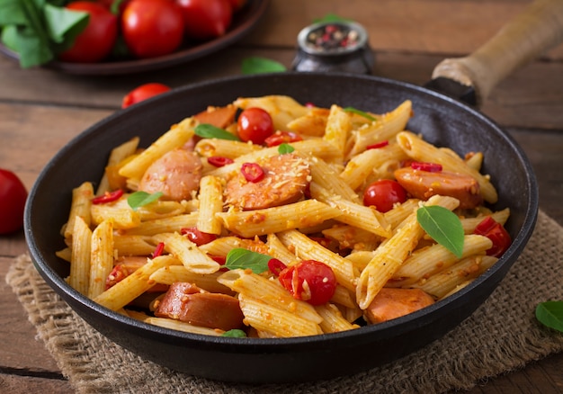 Penne pasta with tomato sauce with sausage, tomatoes, green basil decorated in a frying pan on a wooden table