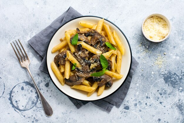 Penne rigate pasta met champignons, parmezaanse kaas en basilicum bladeren in keramische schotel op lichte oude betonnen ondergrond. selectieve focus