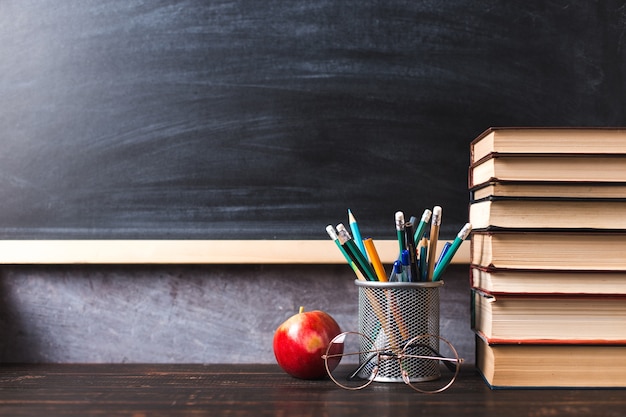 Pennen, appel, potloden, boeken en glazen op tafel, tegen de achtergrond van een schoolbord