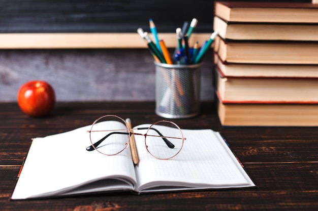 Pennen, appel, potloden, boeken en glazen op tafel, tegen de achtergrond van een schoolbord