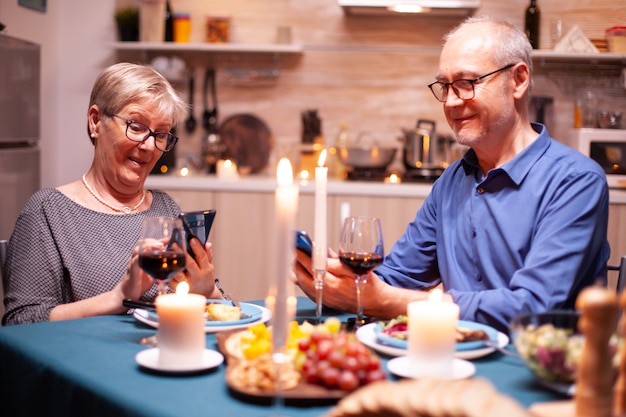 Uomo pensionato che sorride usando il telefono e conversando con la moglie in cucina all'ora di cena. sedersi al tavolo della sala da pranzo, navigare, cercare, usare il telefono, internet,