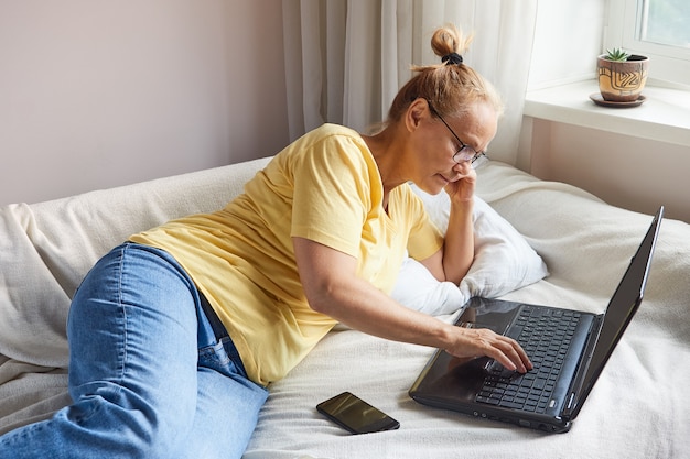 A pensioner with glasses in a yellow shirt uses a laptop while lying on the couch at home. A woman works remotely at a computer, learns online or communicates