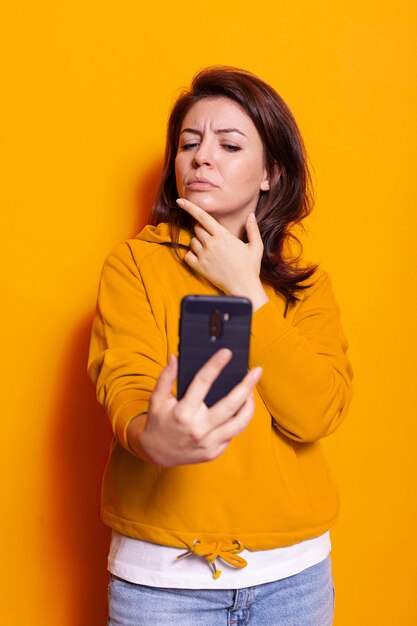 Pensive adult looking at smartphone screen in hand. Woman thinking about technology while using mobile phone with touch screen in studio on camera. Contemplative person with device