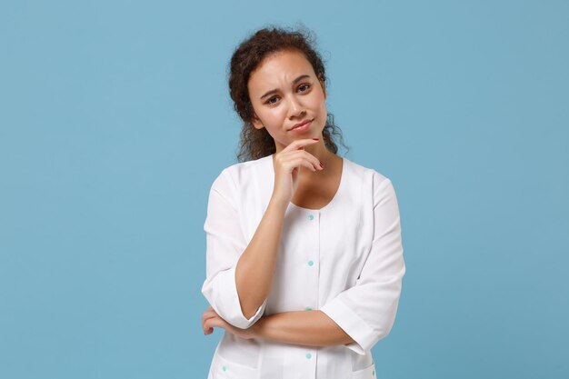 Pensive african american doctor woman isolated on blue wall\
background. female doctor in white medical gown put hand prop up on\
chin. healthcare personnel health medicine concept. mock up copy\
space.