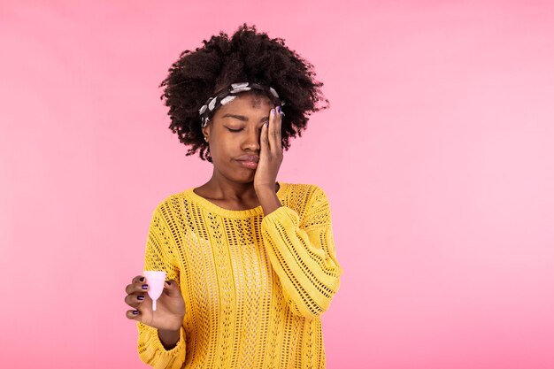 Pensive African American woman holds effective safe affordable menstrual product good protection from leakage chooses reusable menstrual cup thinks how to use it blank empty space on pink wall