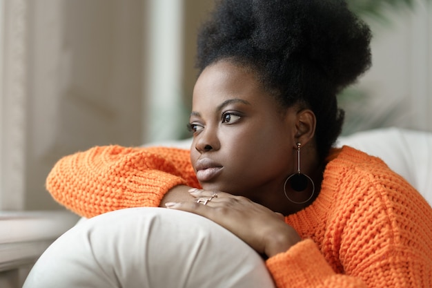 Pensive African millennial woman in orange sweater lying on couch relaxing at home looking at window