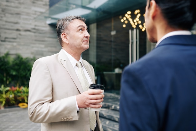 Pensive aged entrepreneur drinking morning coffee with his coworker and looking at office building they are working at