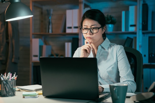 Pensive asian chinese businesswoman at workplace in office in\
front of laptop computer. female designer work creative in late\
time at night. thoughtful girl worker thinking solution on notebook\
pc.