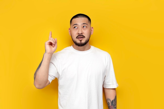 흰색 티셔츠를 입은 잠겨있는 아시아 남자가 손가락을 가리키며 노란색 외진 배경에 대해 생각합니다.
