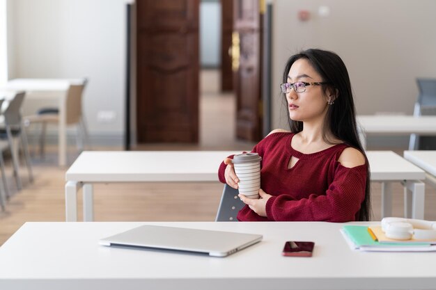 大学図書館で勉強しながらコーヒーを飲み、休んで物思いにふけるアジアの学生の女の子