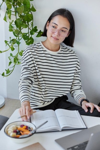 Pensive asian woman with bob hairstyle takes notes in notepad
holds pen looks away being deep in thoughts prepares homework task
writes essay eats delicious meal poses in cozy home interior