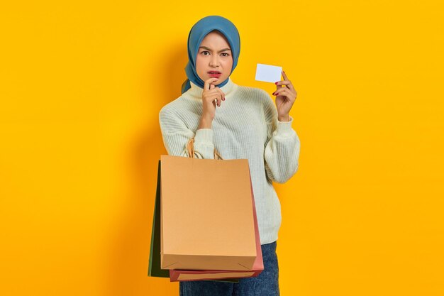 ショッピングバッグとクレジットカードを保持している白いセーターの物思いにふける美しいアジアの女性は、黄色の背景で隔離の何かを考えています