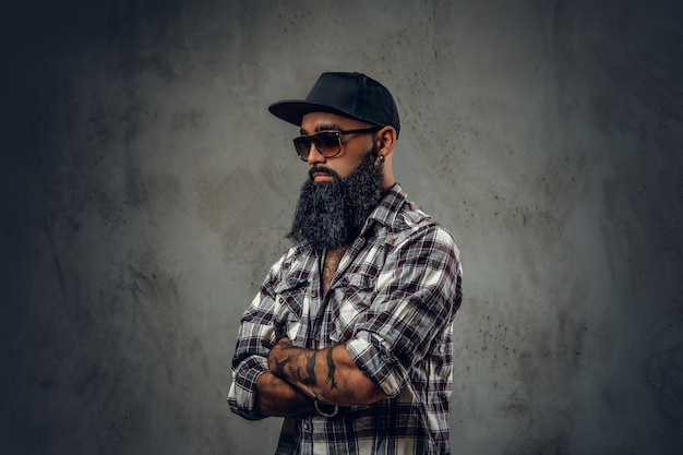 Photo pensive black bearded male dressed in a fleece shirt, sunglasses and baseball cap posing over grey background.