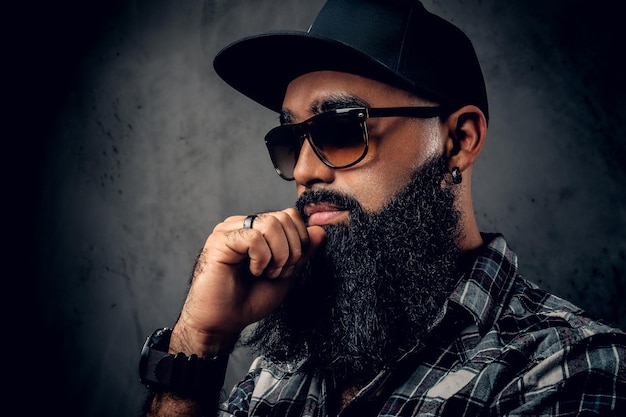 Pensive Black bearded male dressed in a fleece shirt, sunglasses and baseball cap posing over grey background.