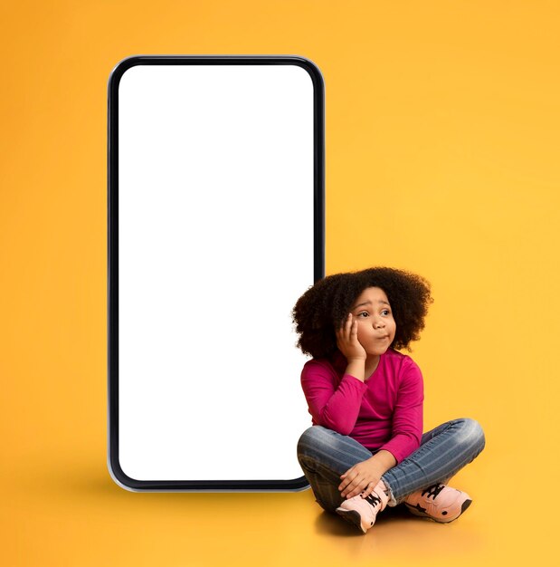 Pensive black little girl sitting next to big blank smartphone