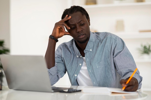 pensive black man taking notes while working with laptop at home office