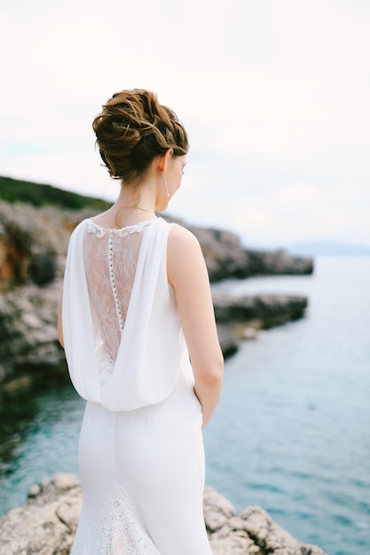 Pensive bride in a lacy white dress stands on a stone shore above the sea back view