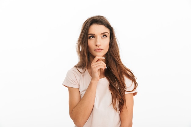 Pensive brunette woman in t-shirt holding her chin and looking away over white wall